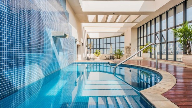 An indoor swimming pool with clear blue water, surrounded by a tiled wall and a wooden deck. - Hb1911 Charme e Estilo no Melhor no Jardins! (São Paulo)