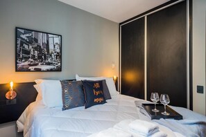 A hotel room with a bed, pillows, a bedside table with glasses, a wall-mounted picture, and a sliding door.