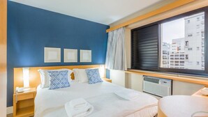 Hotellihuone, jossa on suuri sänky, yöpöytä, ikkuna, jossa on sälekaihtimet, ja näkymä ulkona oleviin rakennuksiin.