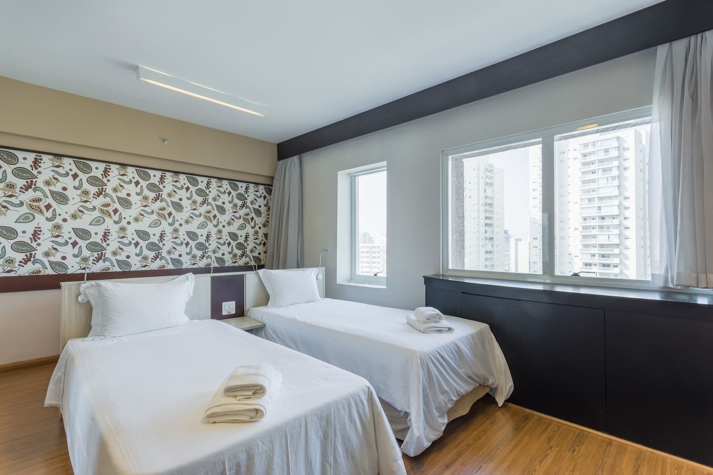 Een hotelkamer met twee bedden, een groot raam met uitzicht op de stad, en behang met een patroon aan de wand.