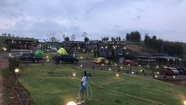 Um espaço ao ar livre com gramado, uma tenda, um edifício com luzes de corda e vários carros estacionados.