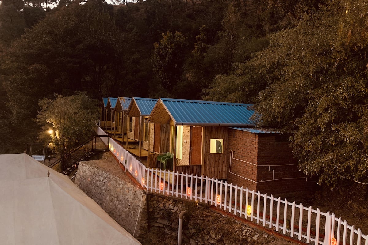A row of wooden cabins with blue roofs, a white picket fence, and a stone wall.
