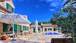 Un patio de piedra con piscina, asientos exteriores y una sombrilla blanca.