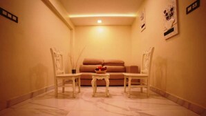 Quarto com piso de mármore, sofá marrom, duas cadeiras brancas e uma pequena mesa com uma fruteira.