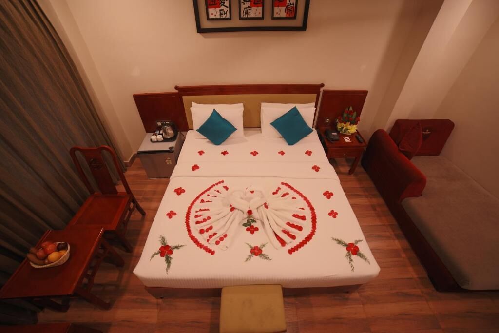 A hotel room with a neatly made bed, a wooden headboard, a red armchair, a small table with a bowl of fruit, and a red sofa.