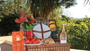 Une table dressée pour un pique-nique, avec un panier en osier contenant du pain, des tomates et une bouteille de boisson, le tout placé à l’extérieur.