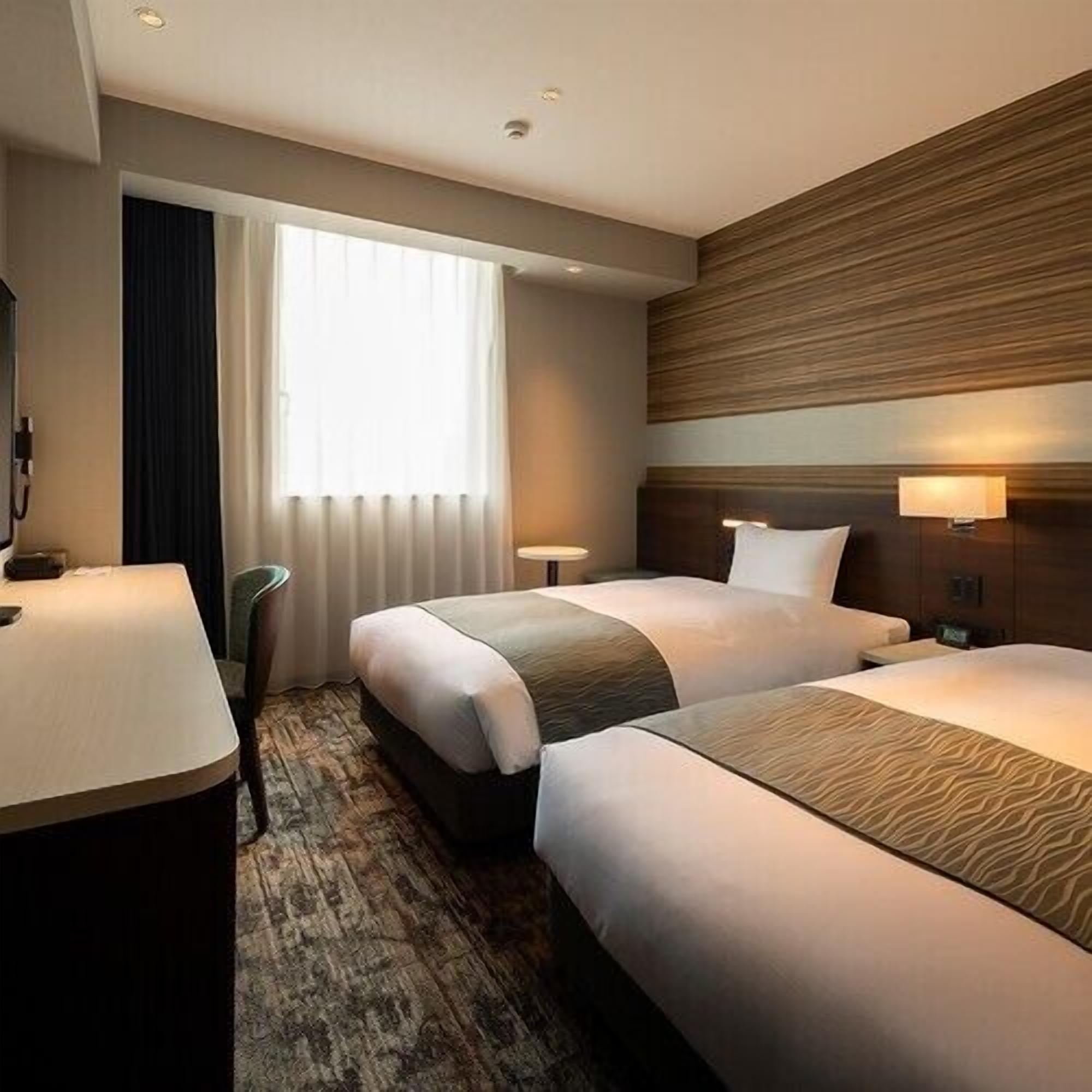 Habitación de hotel con dos camas, un escritorio, una silla, una ventana con cortinas y una lámpara de pared.