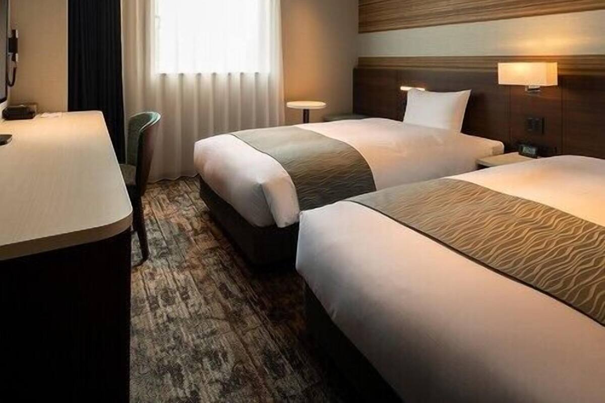 Habitación de hotel con dos camas, un escritorio, una silla, una ventana con cortinas y una lámpara de pared.