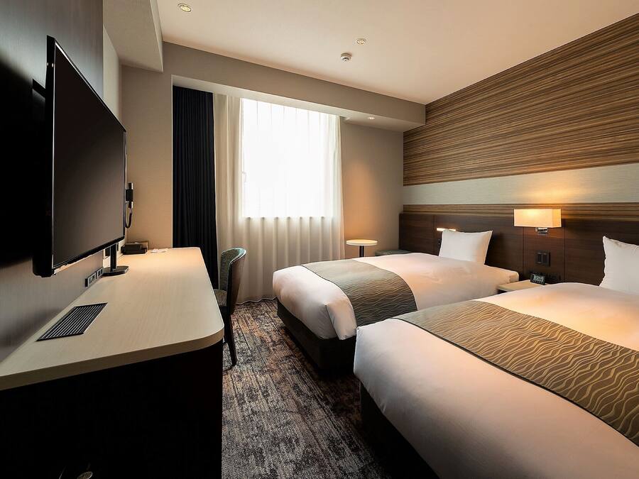 A hotel room with two beds, a desk, a TV, and a window with curtains.