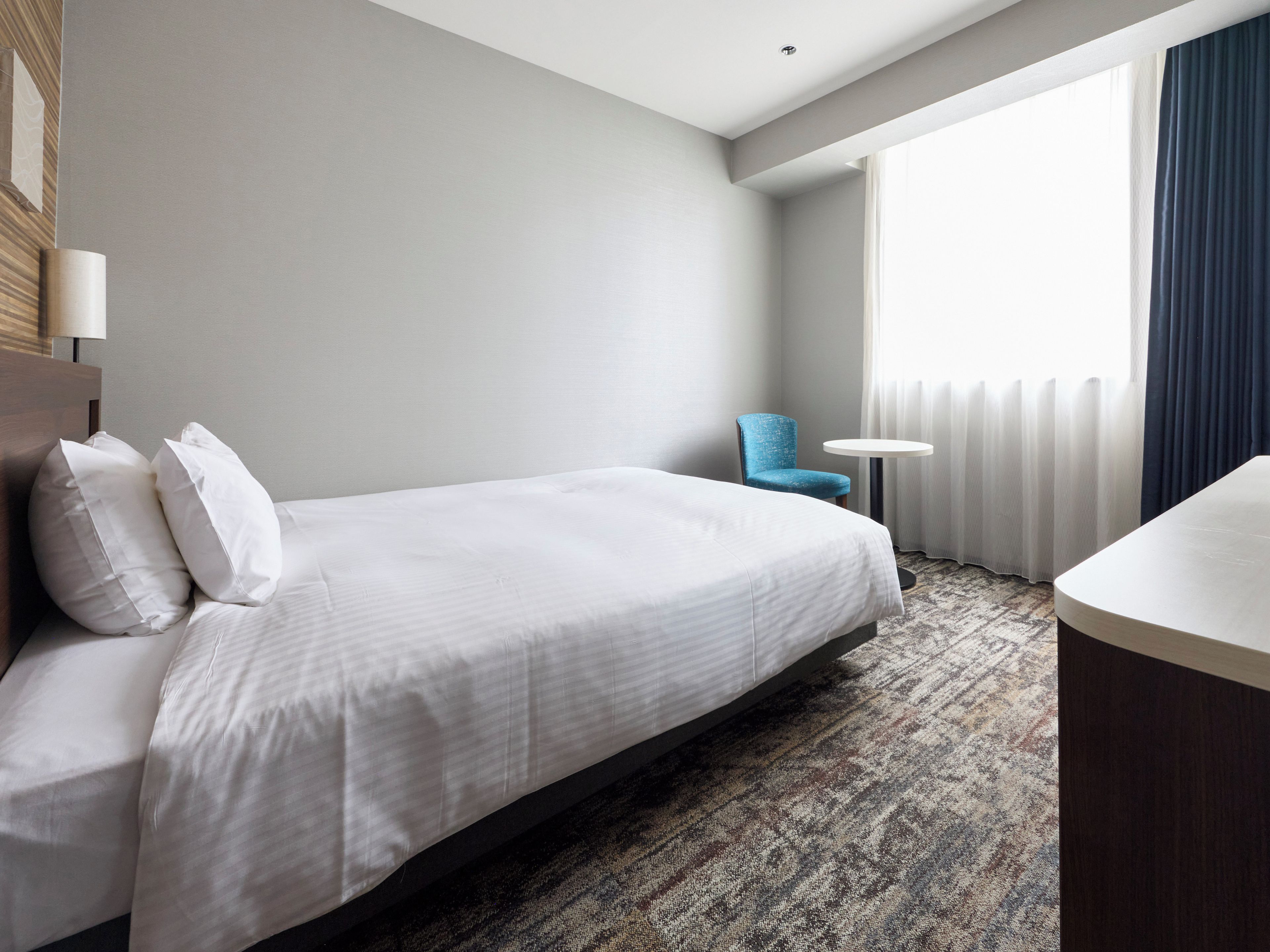 Une chambre d’hôtel avec un lit, une chaise, une petite table ronde et une fenêtre avec des rideaux.