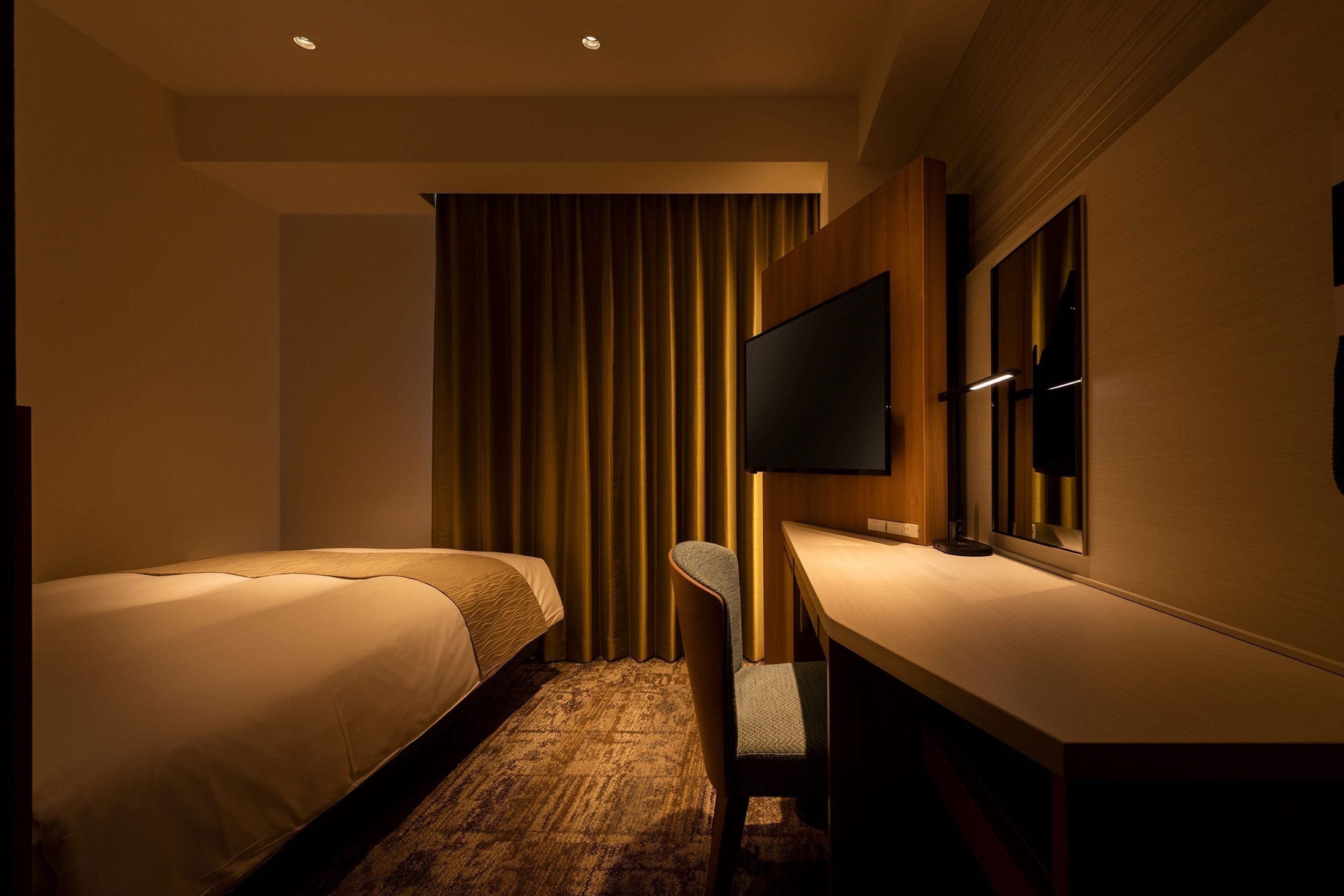 A hotel room with two beds, a desk, a chair, a flat-screen TV mounted on the wall, and a large window with curtains.