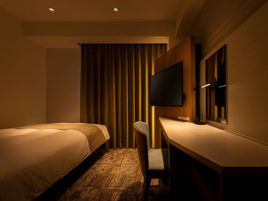 A hotel room with two beds, a desk, a chair, a flat-screen TV mounted on the wall, and a large window with curtains.