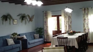 A living room with a blue sofa, a dining table with chairs, and a large window with floral curtains. - Villa Flor Campos (Cork Oak) (Vila Nova de Cerveira)