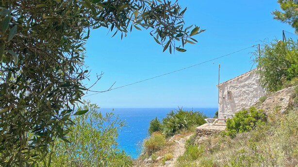 Un sentier côtier bordé d’un bâtiment en pierre, d’oliviers et offrant une vue dégagée sur la mer.