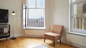Ein Zimmer mit einem großen Bogenfenster, einer schwarzen Stehlampe, einem rosafarbenen Sessel und einem Fernseher auf einem Podest.