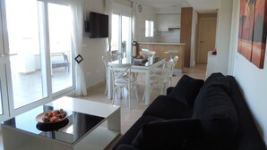 Uma sala de estar moderna com um sofá preto, uma mesa de centro de vidro e uma área de jantar com cadeiras brancas e uma mesa.