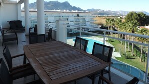 Varanda com mesa e cadeiras de madeira, com vista para uma piscina e uma paisagem montanhosa.