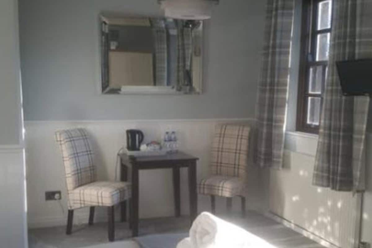 A small, cozy room with a bed, two chairs, a table, a mirror, and a window with curtains.