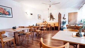 A spacious dining area with wooden tables and chairs, a ceiling fan, and various wall decorations including framed artwork and plants. - Hotel Garni Rank (Bad Berka)