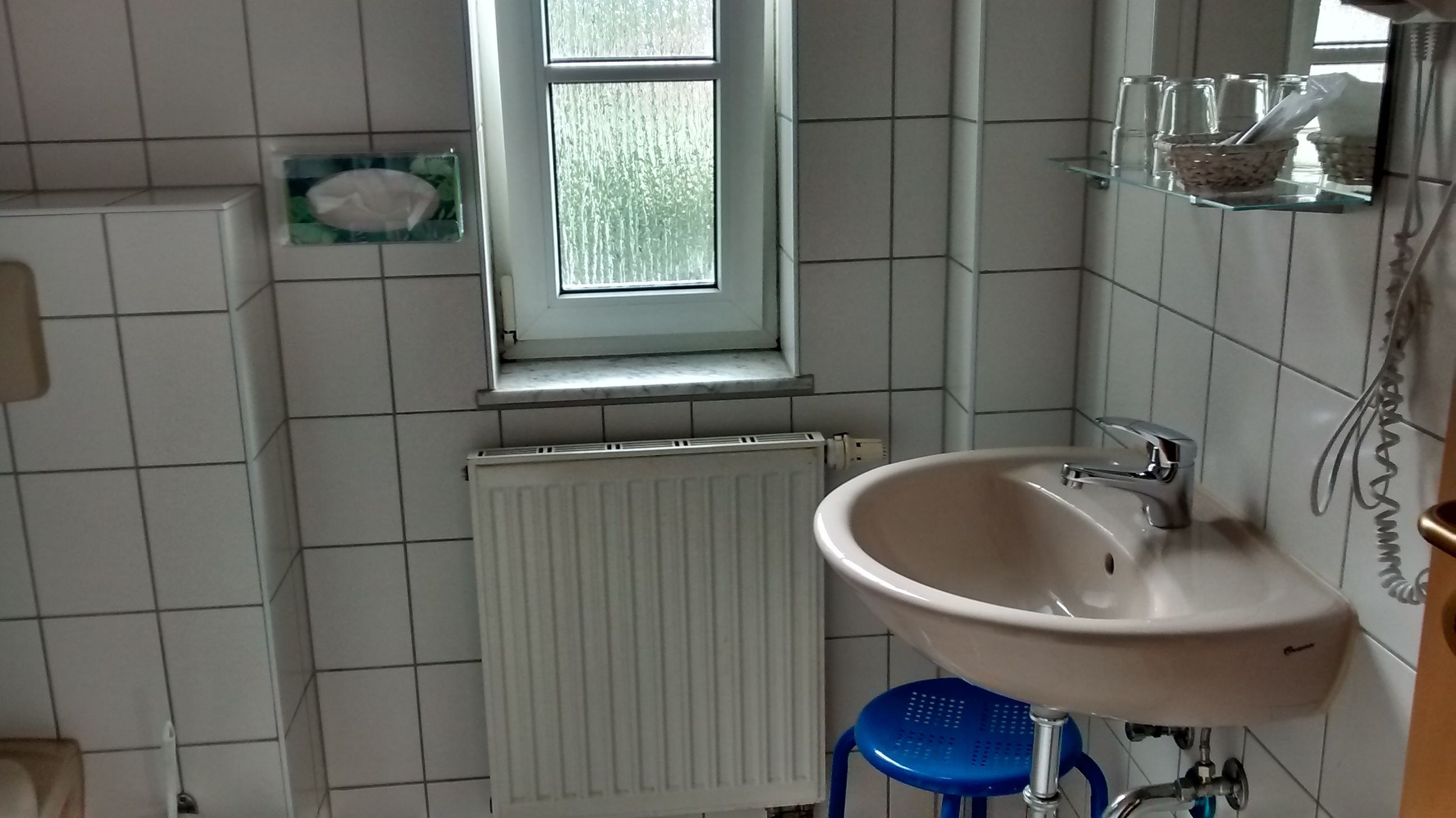 Ein Badezimmer mit einem weißen Waschbecken, einem Fenster, einem Heizkörper und einem blauen Hocker.