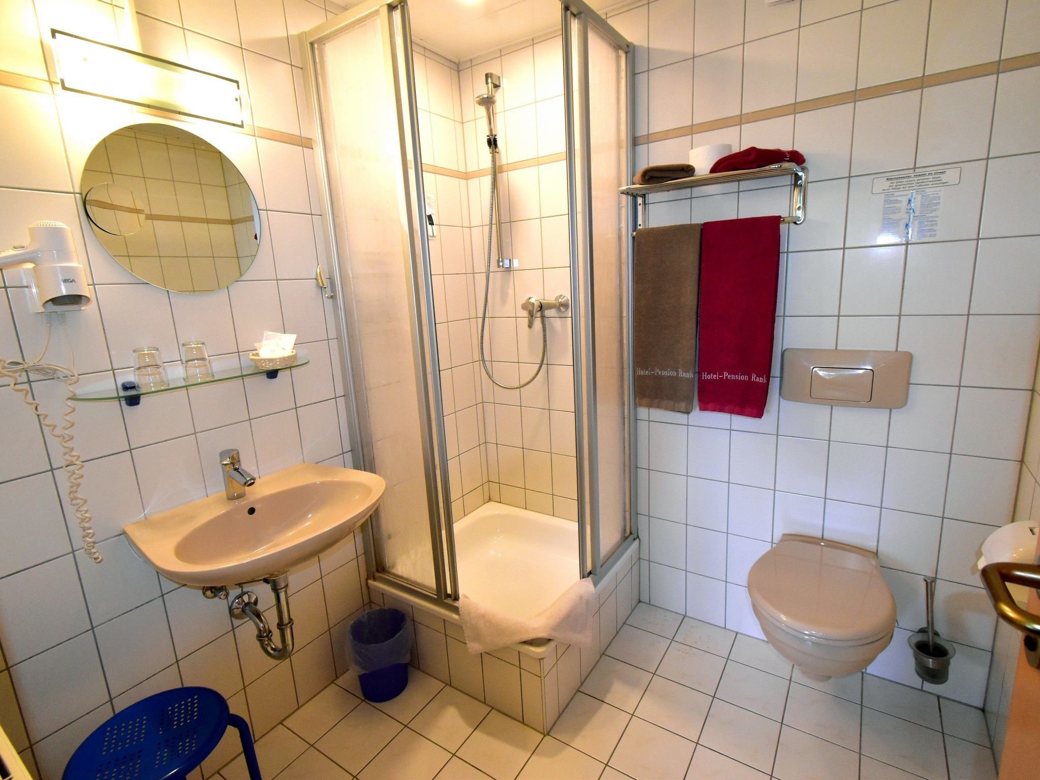 A bathroom with a sink, toilet, and shower. There is a towel rack with towels, a mirror, and a trash bin.