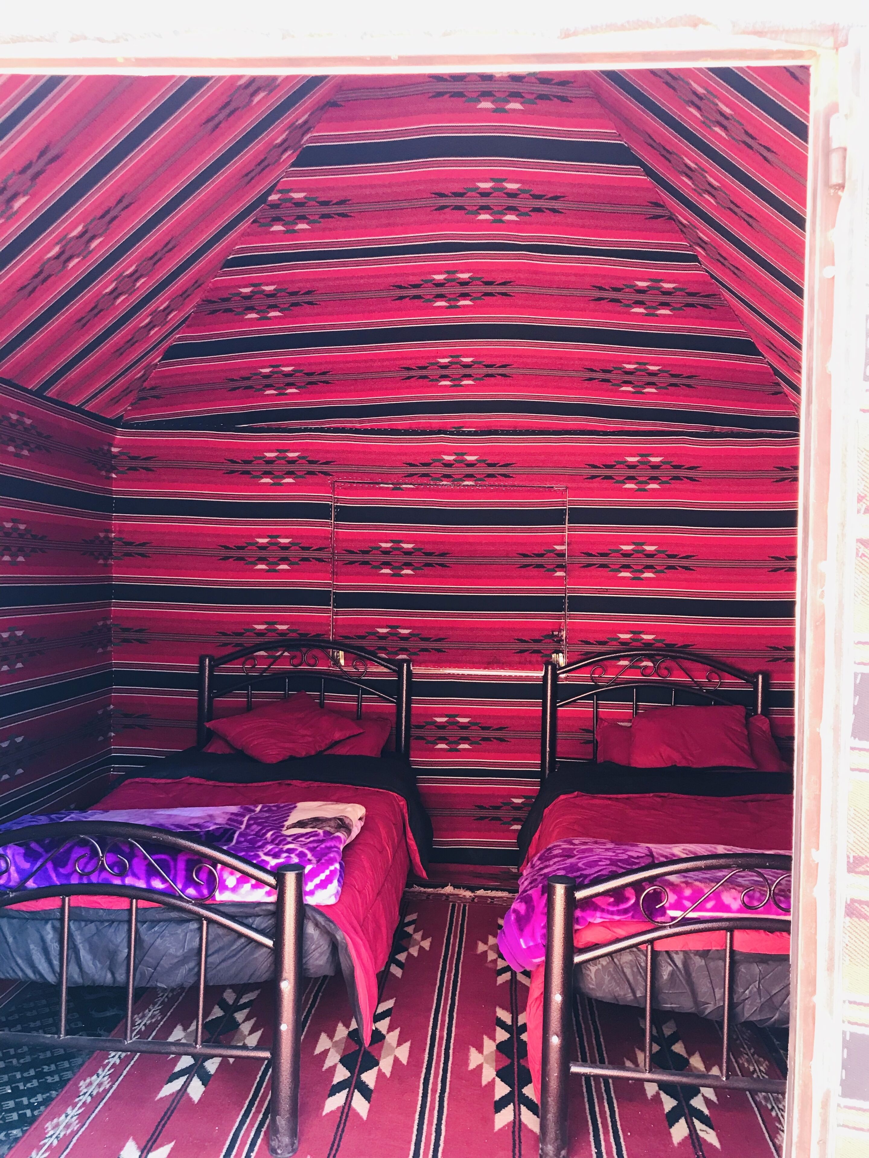 A room with two beds, each with a metal frame and red bedding. The walls and floor have a striped pattern.