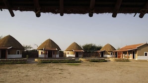 A row of thatched-roof cottages with a sandy ground in between. - Rann Visamo Village Resort (Bhuj)