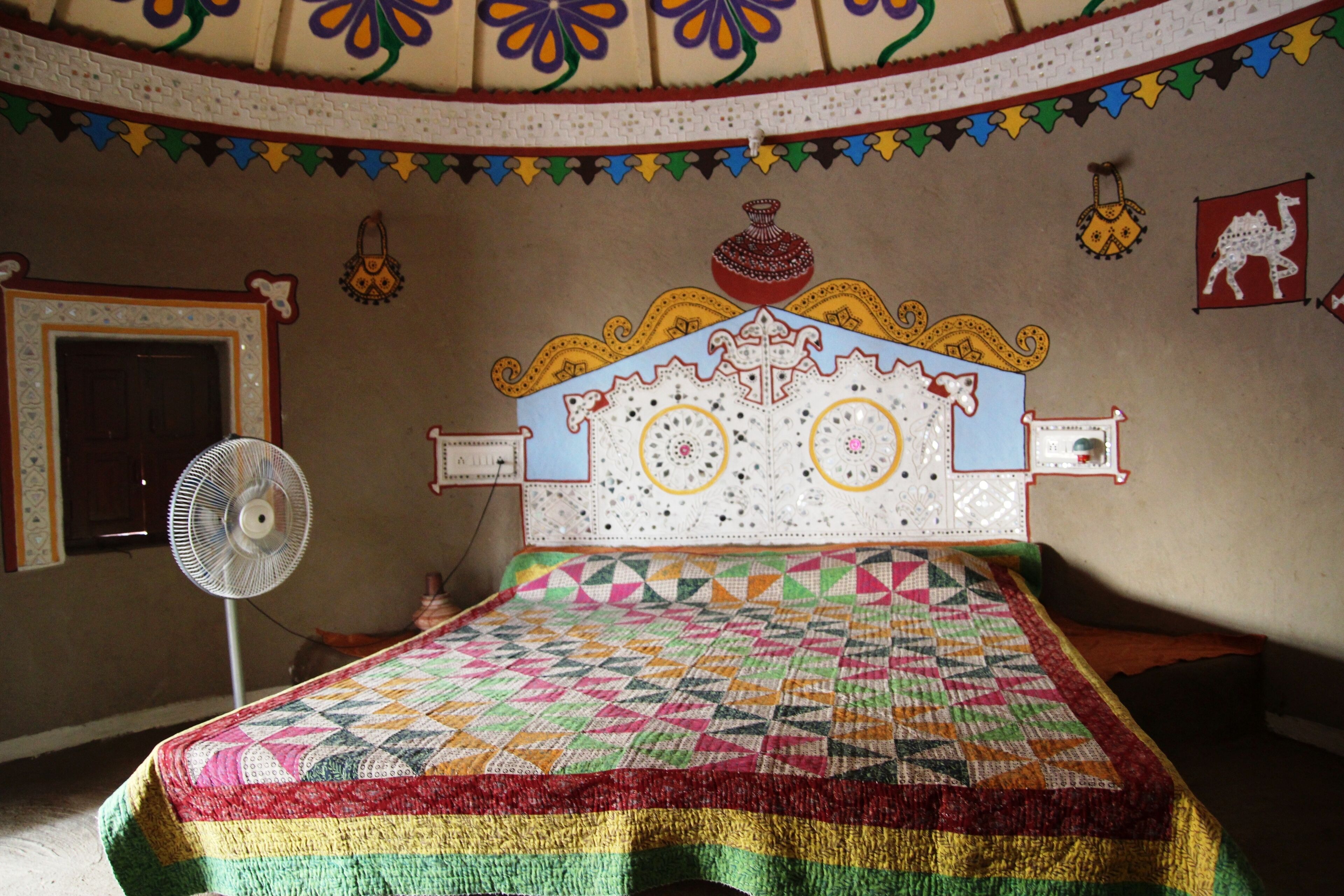 A room with a bed, a fan, and a wall with decorative patterns and a camel flag.