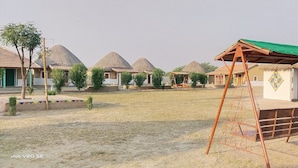 A rural village with thatched-roof houses, a swing set, and a small fenced area. - Rann Visamo Village Resort (Bhuj)