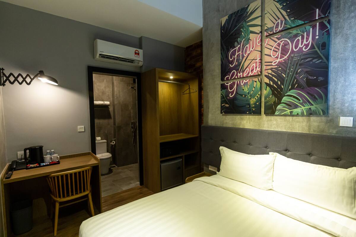 Quarto de hotel com cama, escrivaninha com cadeira, guarda-roupa e ar condicionado.