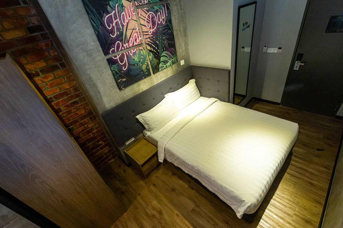 Quarto com cama, mesa de cabeceira, piso de madeira, parede de tijolos e um pôster na parede.