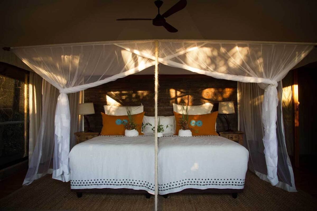 Cama doble con ropa de cama blanca, dos mesitas de noche con lámparas, y una cama con dosel y cortinas translúcidas.