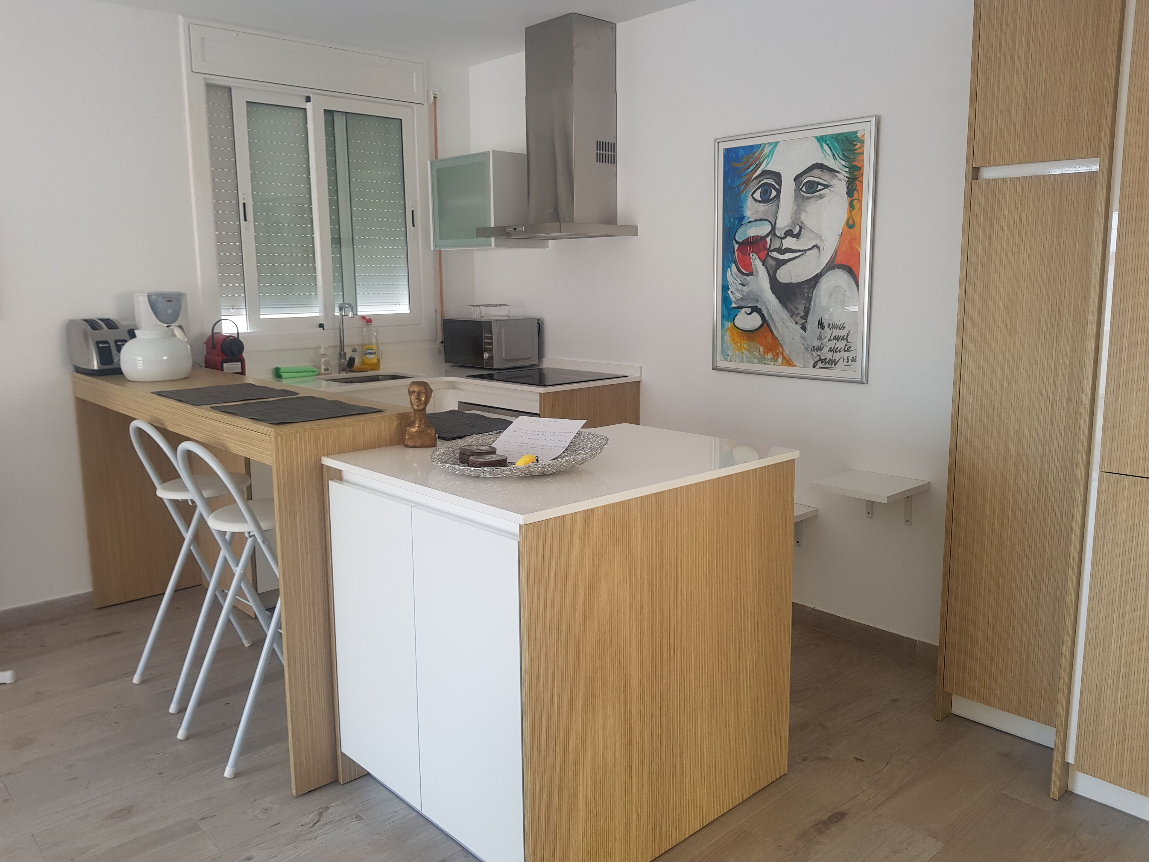 A compact kitchen with a microwave, toaster, and sink. There's a small island with a paper and a banana. A chair is next to the island.