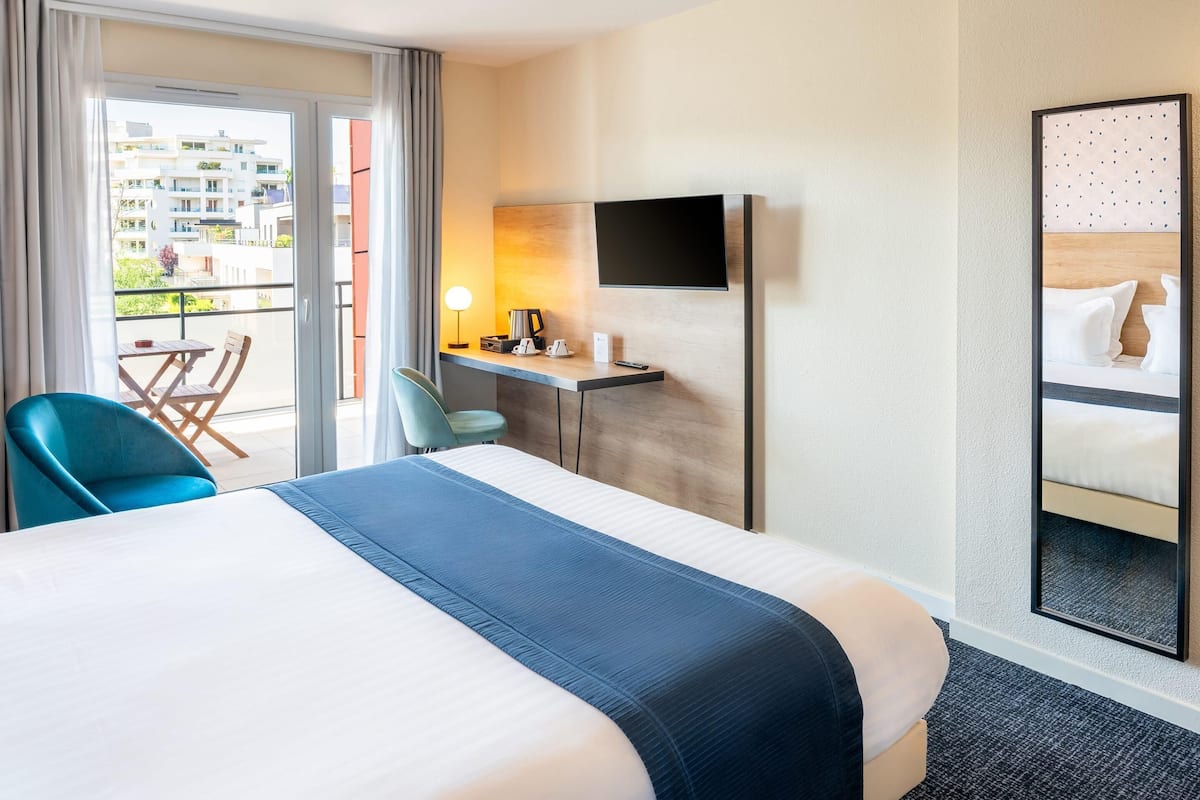 Une chambre d’hôtel avec un grand lit, un bureau avec une chaise, un téléviseur fixé au mur et un balcon donnant sur d’autres immeubles.