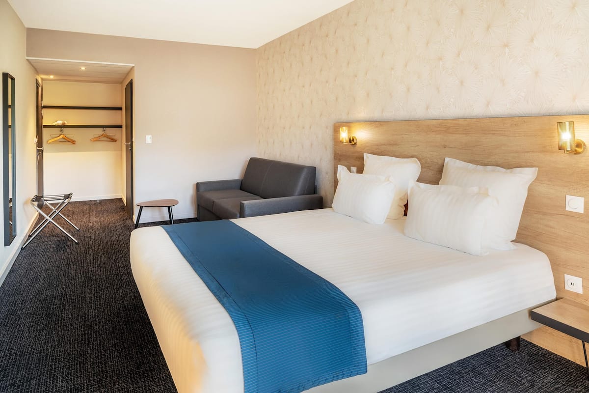 Une chambre d’hôtel avec un grand lit, un fauteuil gris, une petite table et une armoire contenant des vêtements suspendus.