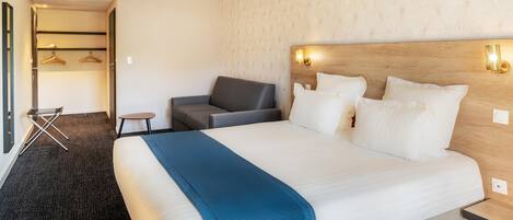 Una habitación de hotel con una cama grande, un sillón gris, una mesita pequeña y un armario con ropa colgada.