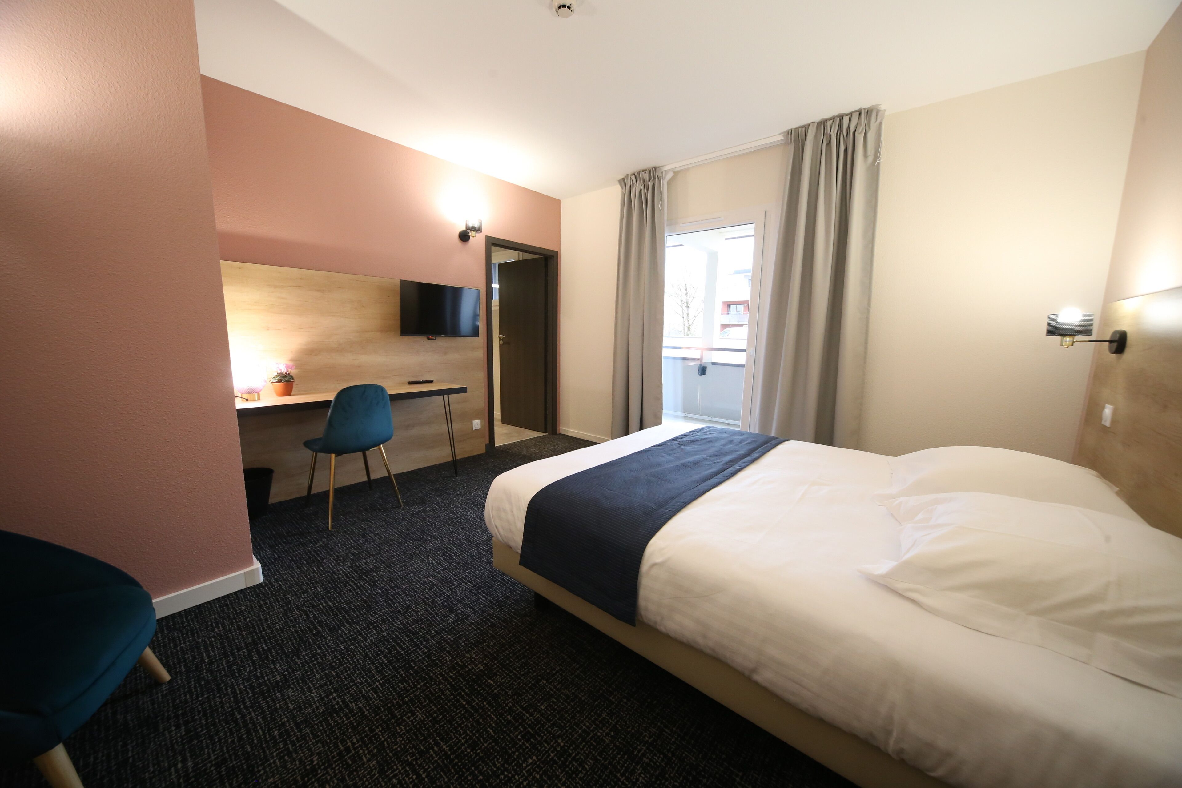 A hotel room with a bed, a desk with a chair, a television, and a window with curtains.