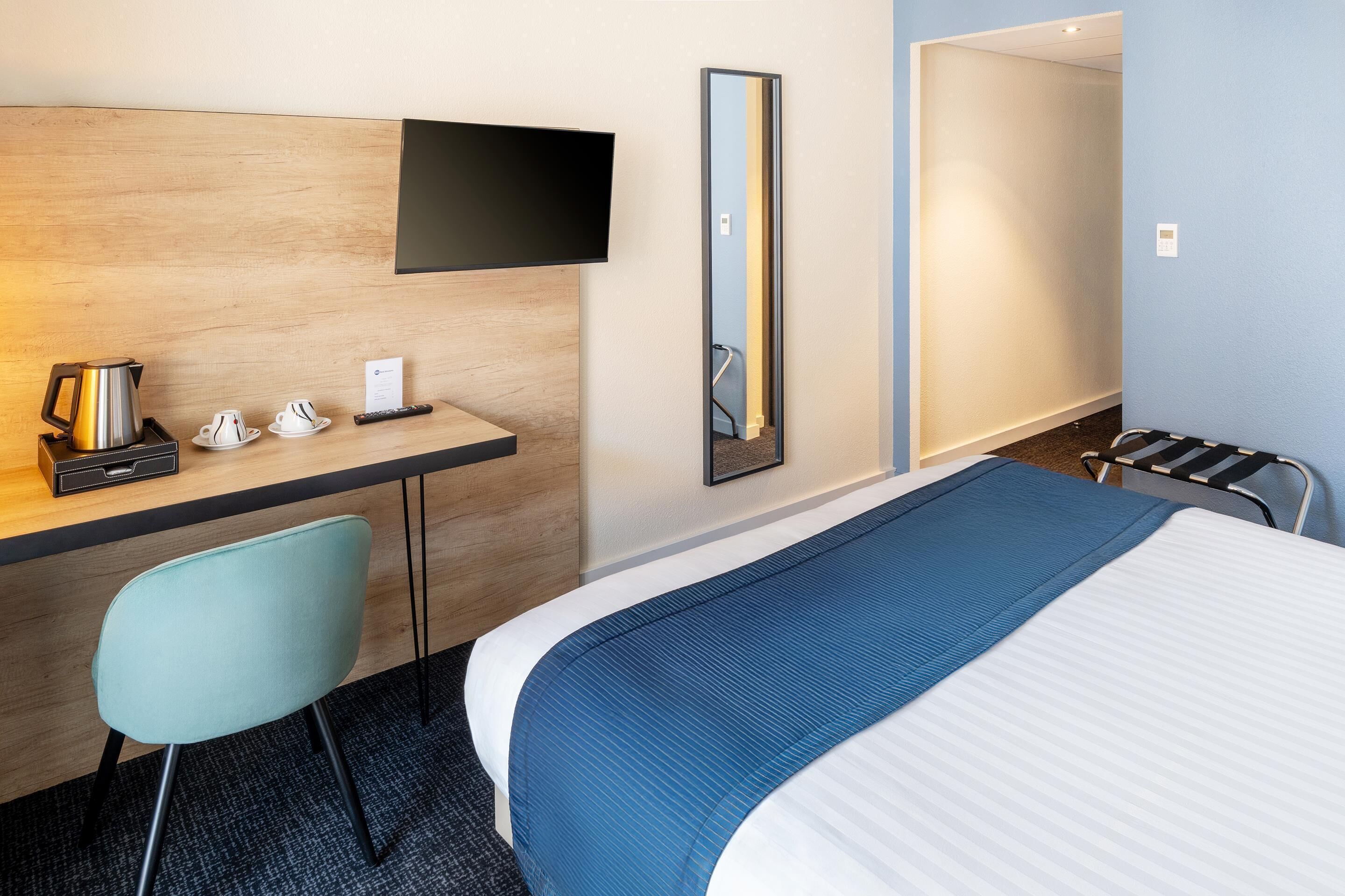 Een hotelkamer met een bed, een bureau met stoel, een aan de muur bevestigde televisie en een spiegel.