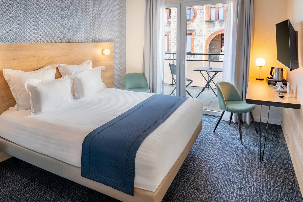 Une chambre d’hôtel avec un grand lit, un bureau avec une chaise, une télévision et un balcon équipé d’une table et de chaises.