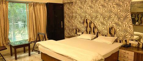 A bedroom with a large bed, a desk, and a chair. There is a window with curtains, a door, and a patterned wall.