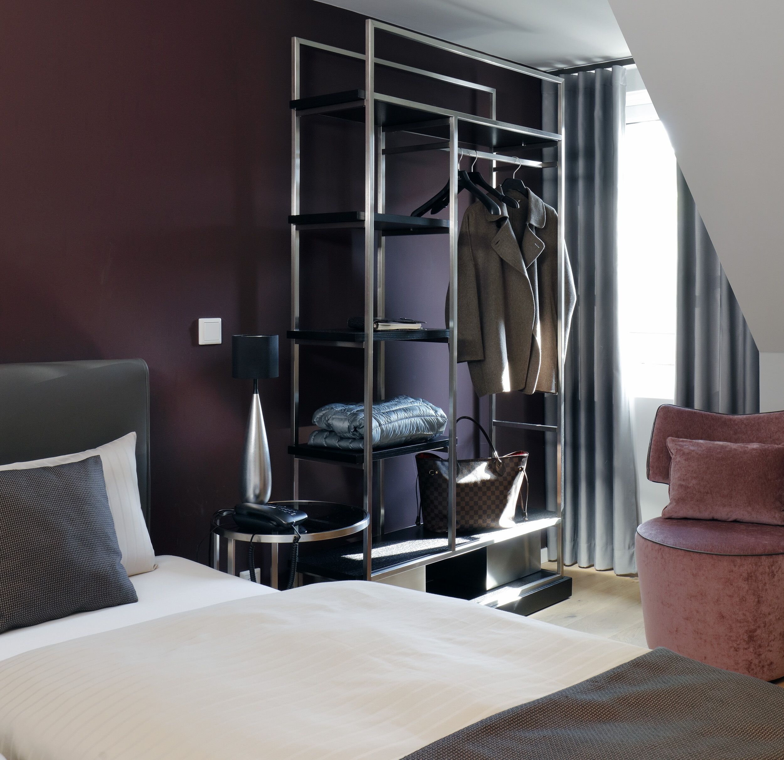 A modern hotel room with a large bed, a wardrobe with hanging clothes, a desk with a lamp, and a pink armchair.