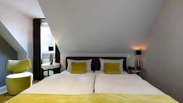 Quarto de hotel com uma cama grande, duas almofadas, uma mesa de cabeceira com abajur, uma cadeira e uma janela com cortinas.