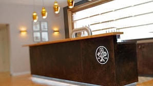 A reception desk with a logo, pendant lights, and a window with blinds. - Patentkrug Design Hotel (Oldenburg)