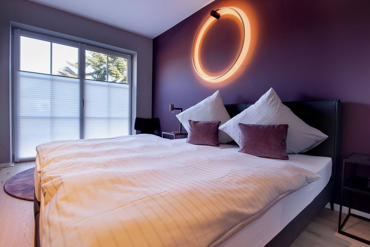 A modern bedroom with a large bed, a circular light fixture, and a view of the outdoors through a window.