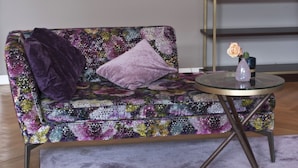 A sofa with a floral pattern, a purple throw blanket, and a vase with a single rose on a glass table. - Patentkrug Design Hotel (Oldenburg)