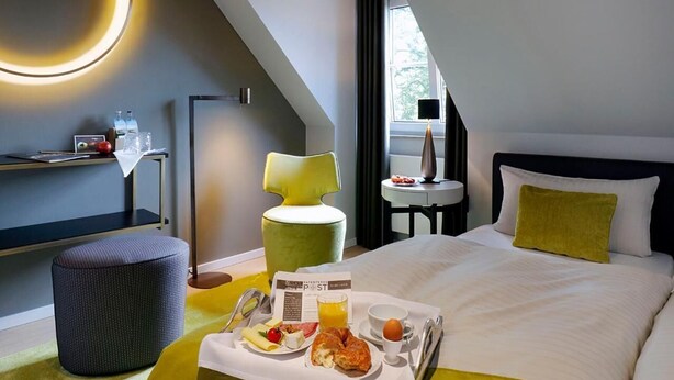 A hotel room with a bed, a dining table with breakfast, a chair, a lamp, and a window with curtains. - Patentkrug Design Hotel (Oldenburg)