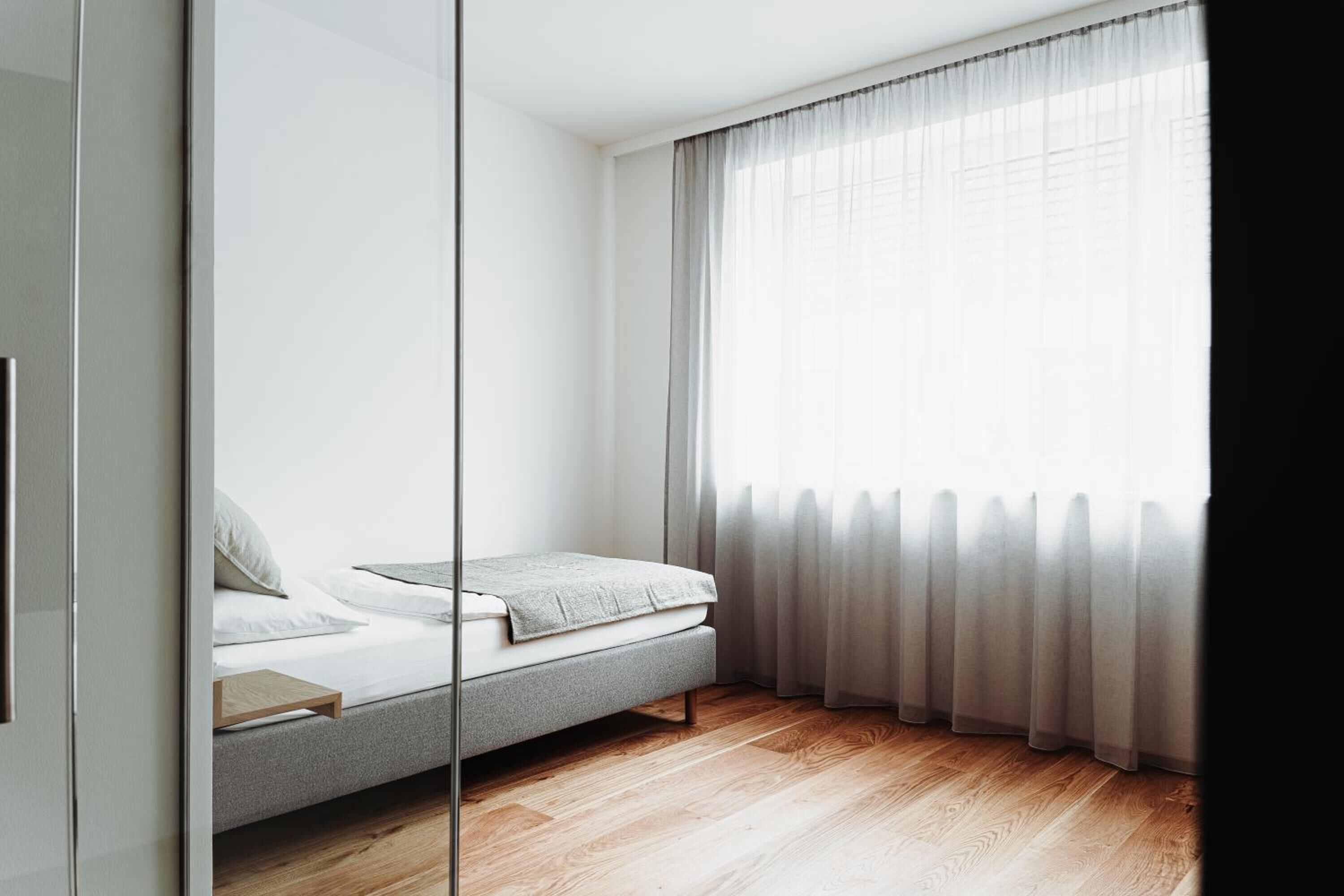 A modern bedroom with a single bed, a wooden floor, and sheer curtains.