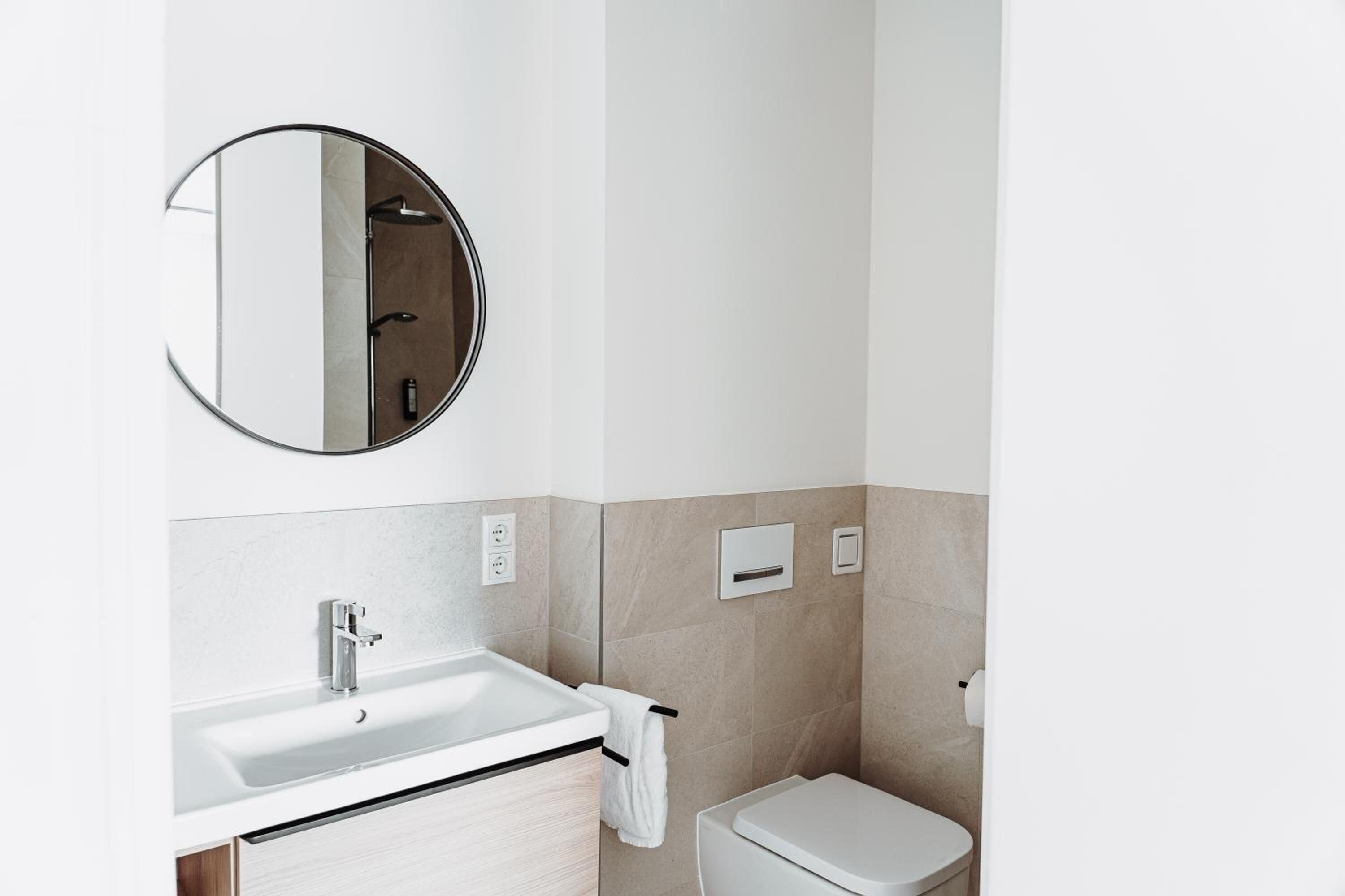 Een moderne badkamer met een wit wastafel, een ronde spiegel, een toilet en beige tegels.
