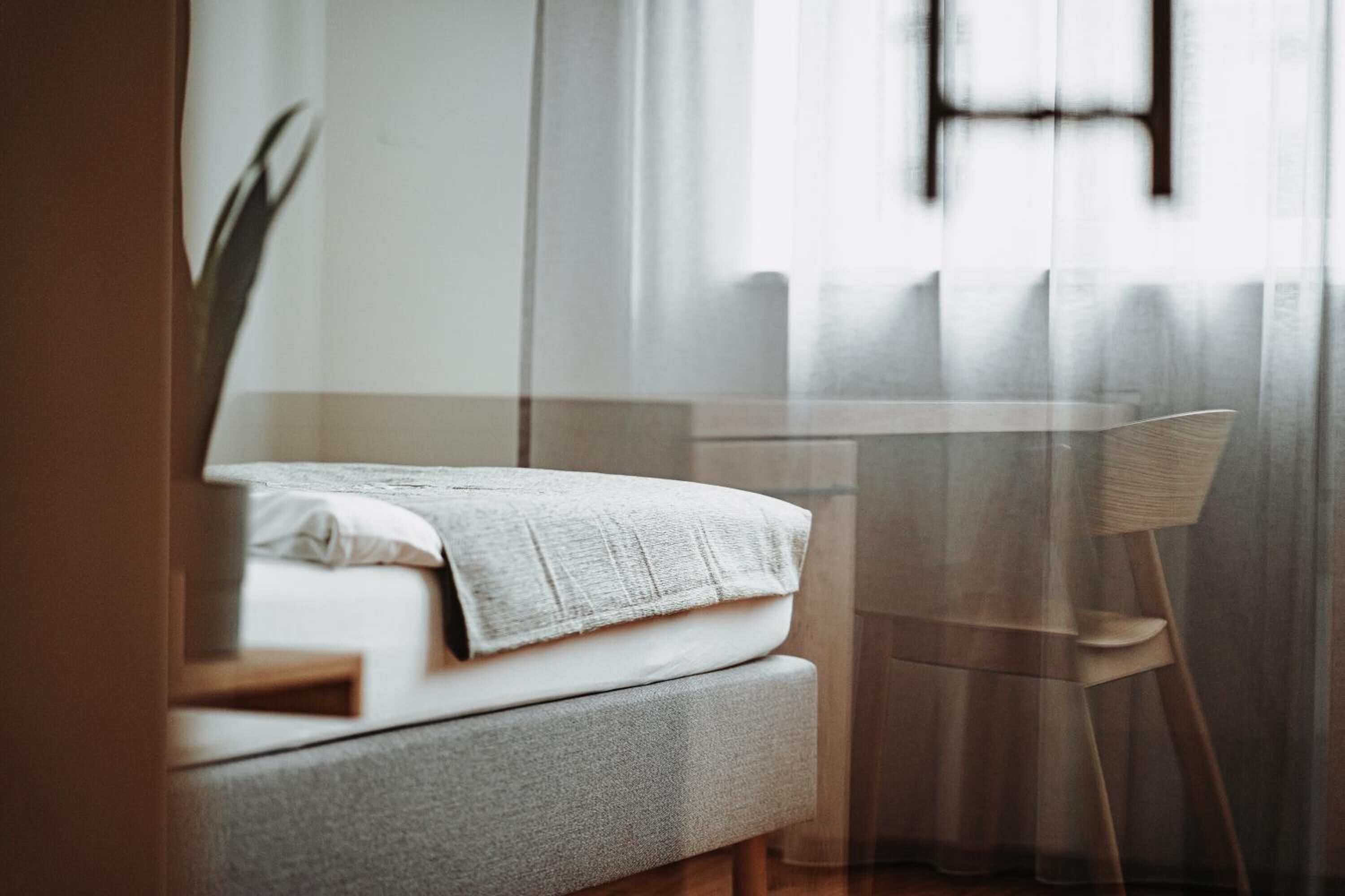 Un lit bien fait, avec un oreiller blanc et une couverture grise, une table en verre transparent et une chaise en bois, dans une pièce où sont accrochées des rideaux légers.