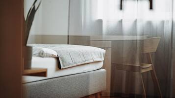 Un lit bien fait, avec un oreiller blanc et une couverture grise, une table en verre transparent et une chaise en bois, dans une pièce où sont accrochées des rideaux légers.